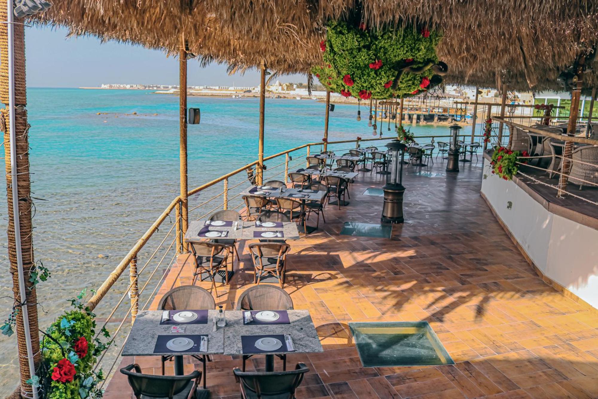 라구나 비치 럭셔리 리조트 앤 스파 마나마 외부 사진 The photo shows a seaside restaurant with a spacious outdoor dining area. It features several tables set with plates and utensils, surrounded by lush greenery and decorative plants. The flooring is made of brown tiles, and there are thatched roofs pr