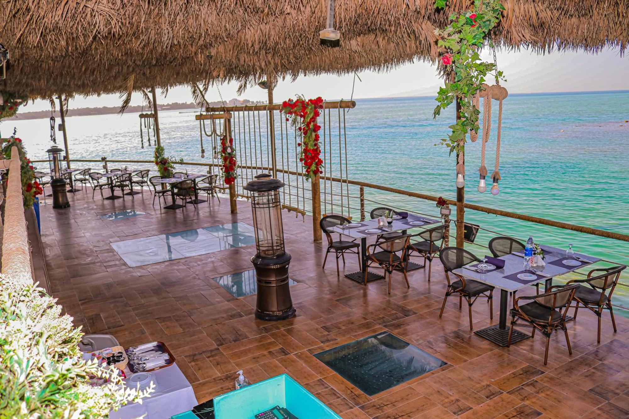라구나 비치 럭셔리 리조트 앤 스파 마나마 외부 사진 The photo depicts a coastal restaurant or dining area with an open-air design. It features a wooden floor and tables set for dining, surrounded by chairs. The space is partially covered by a thatched roof, providing shade while allowing for a view of