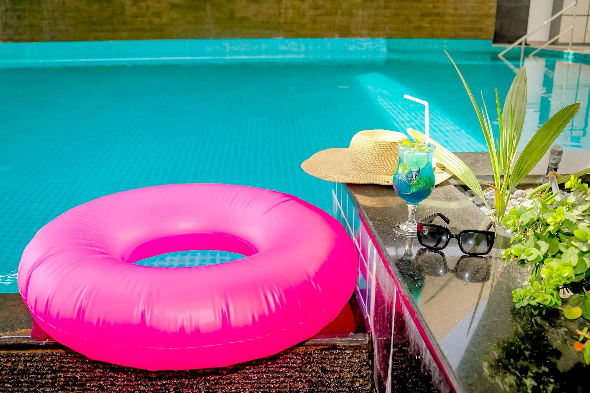 라구나 비치 럭셔리 리조트 앤 스파 마나마 외부 사진 The photo shows a vibrant and inviting scene by a swimming pool. In the foreground, there is a large pink inflatable float and a stylish straw hat. Next to the float, a refreshing cocktail in a glass is displayed, complete with a straw and garnishes.
