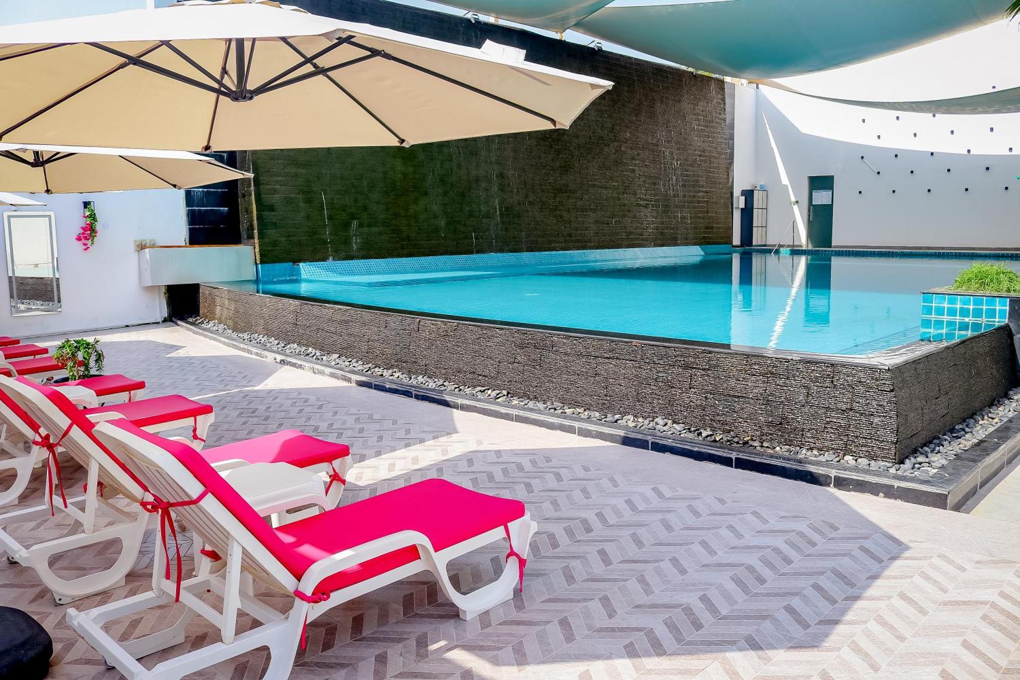 라구나 비치 럭셔리 리조트 앤 스파 마나마 외부 사진 The photo shows a pool area featuring a rectangular swimming pool with clear blue water. Surrounding the pool are light-colored lounge chairs with pink cushions, positioned under large umbrellas for shade. The pool is bordered by a textured wall, and