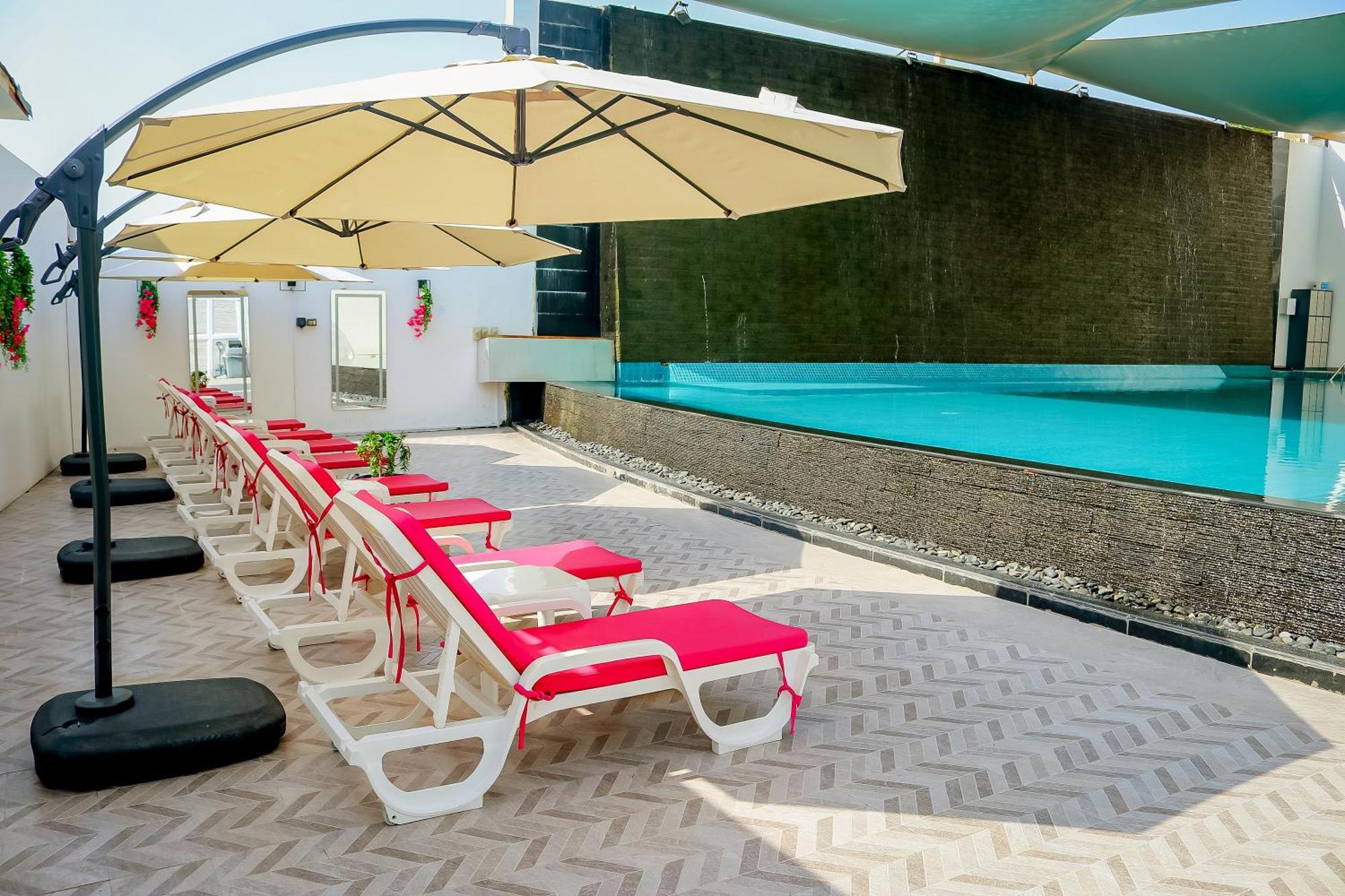 라구나 비치 럭셔리 리조트 앤 스파 마나마 외부 사진 The photo shows a sunny poolside area with several lounge chairs arranged in a row. Each chair features red cushions and is positioned under large, beige umbrellas for shade. In the background, there is a beautiful swimming pool with clear blue water