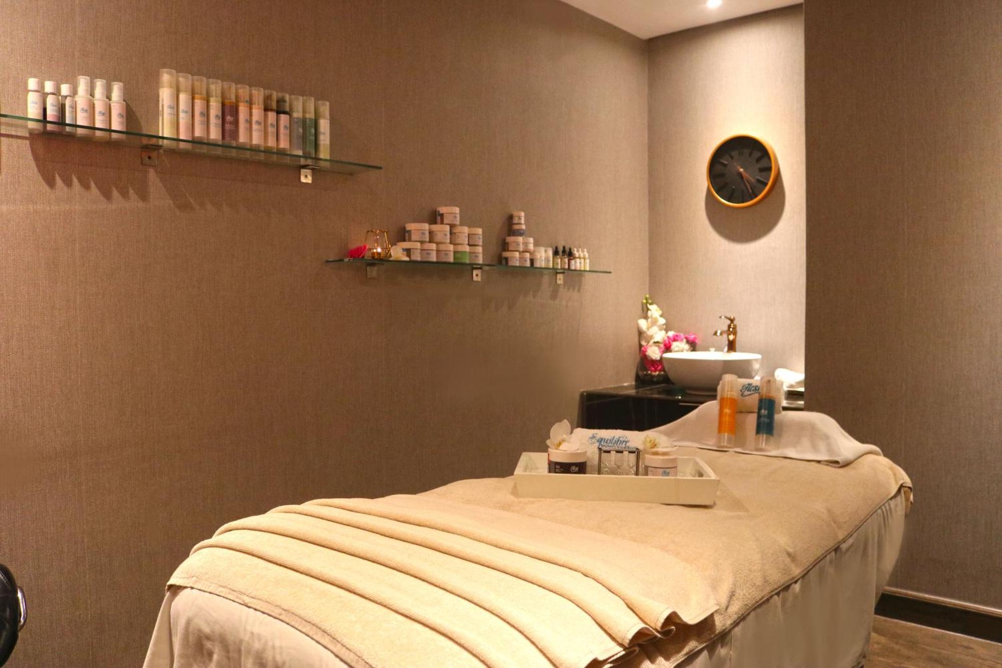 라구나 비치 럭셔리 리조트 앤 스파 마나마 외부 사진 The photo shows a spa treatment room. There is a massage table draped with a light-colored blanket, creating a relaxing atmosphere. On the wall, there are shelves displaying various skincare products in bottles and jars. A clock is mounted on the wal
