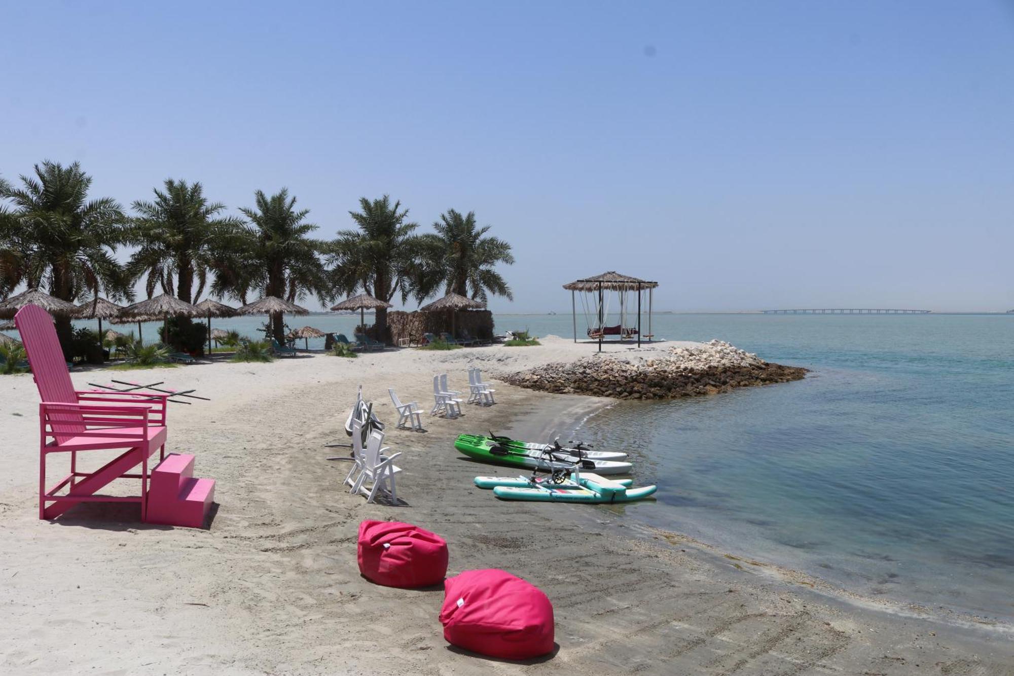 라구나 비치 럭셔리 리조트 앤 스파 마나마 외부 사진 The photo depicts a serene beach scene with clear blue water and a sunny sky. In the foreground, there are colorful pink bean bags and a large pink chair on the sand. A few white chairs and loungers are positioned near the water's edge. To the side, 