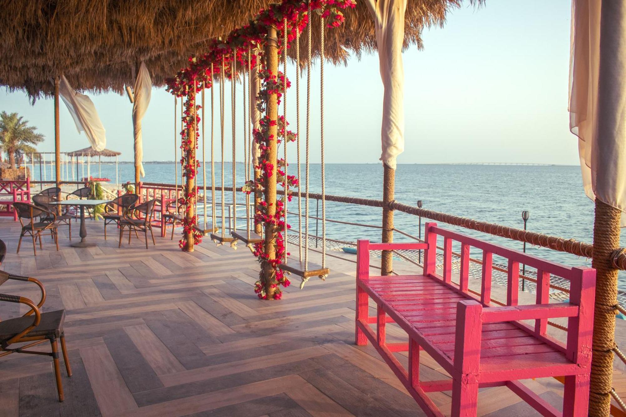 라구나 비치 럭셔리 리조트 앤 스파 마나마 외부 사진 The photo depicts a scenic outdoor area by the water, with a wooden deck and a thatched roof. Two swings adorned with flowers are hanging from the ceiling, providing a charming touch to the space. In the foreground, there is a bright pink bench, addi