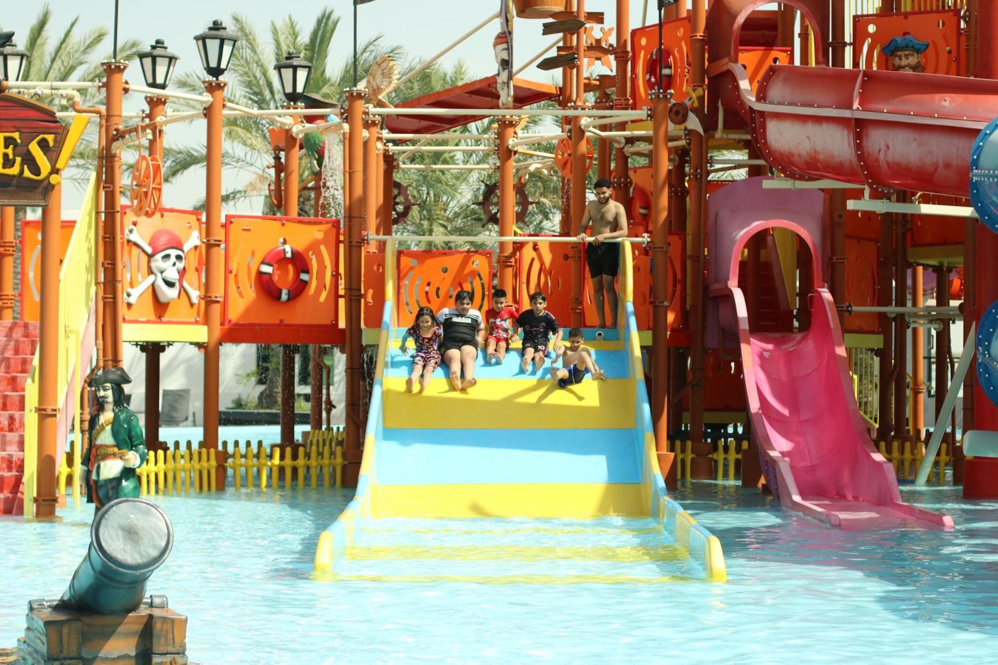 라구나 비치 럭셔리 리조트 앤 스파 마나마 외부 사진 The photo shows a colorful water park scene featuring a play area with multiple slides. There are children enjoying themselves, sliding down bright yellow and pink slides into a pool of water. The structure has pirate-themed decorations, including a 