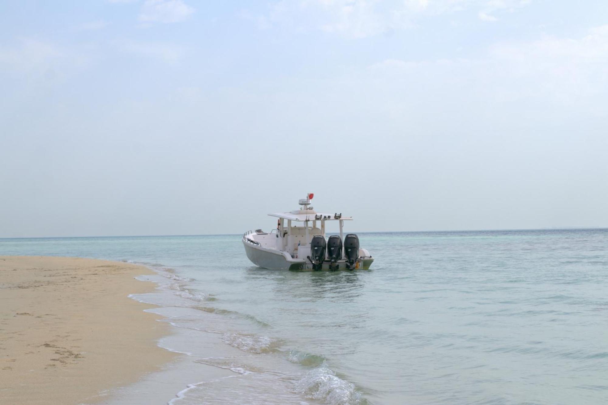 라구나 비치 럭셔리 리조트 앤 스파 마나마 외부 사진 The photo depicts a small boat anchored near a sandy beach, partially submerged in calm, clear water. The boat is white and features three outboard motors on its stern. In the background, the sea stretches out toward the horizon under a light, cloudy