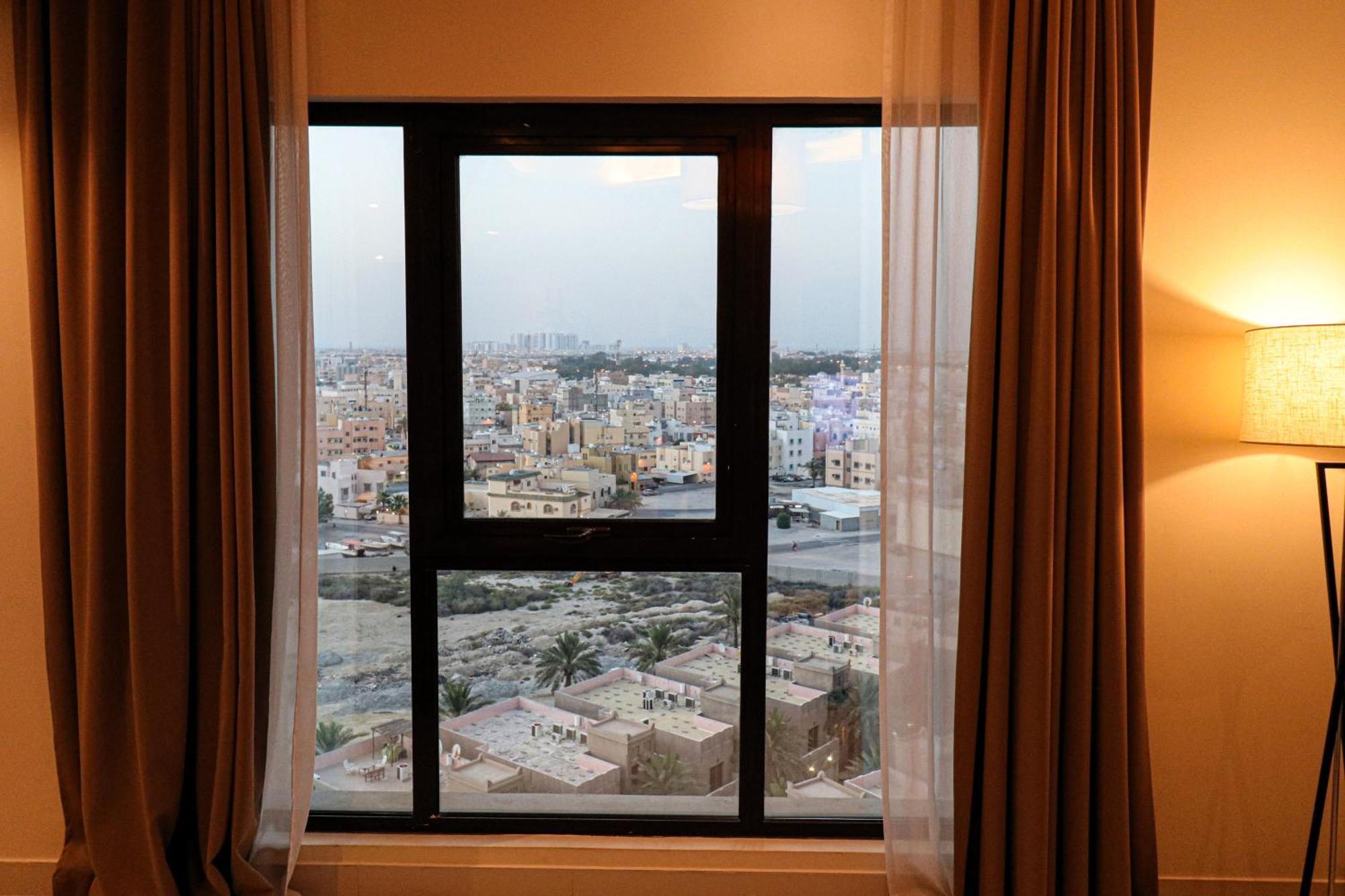 라구나 비치 럭셔리 리조트 앤 스파 마나마 외부 사진 The photo shows a window with a black frame and sheer curtains. Through the window, there is a view of a cityscape, displaying a densely populated area with various buildings and rooftops. The sky appears slightly overcast, suggesting a muted lightin