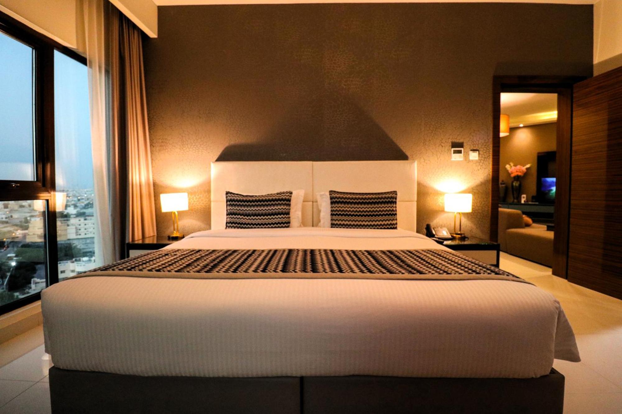 라구나 비치 럭셔리 리조트 앤 스파 마나마 외부 사진 The photo depicts a modern hotel room featuring a large bed with a white comforter and two decorative pillows. The headboard is light-colored, and the walls are painted a dark hue. On either side of the bed, there are bedside tables with lamps. The r