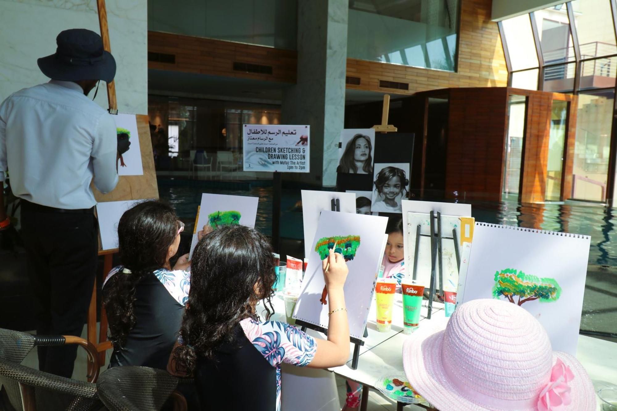 라구나 비치 럭셔리 리조트 앤 스파 마나마 외부 사진 The photo shows a group of children and an adult engaged in an art activity near a pool area. The children are seated at a table, painting on canvases or paper, with a focus on trees, as indicated by the green paint they are using. An adult, wearing 