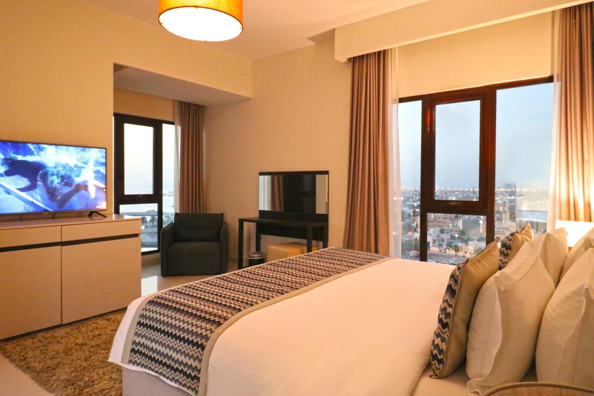 라구나 비치 럭셔리 리조트 앤 스파 마나마 외부 사진 The photo shows a modern hotel room or suite. It features a neatly made bed with decorative pillows in the foreground. To the left, there is a TV on a cabinet, and further back, a comfortable chair is situated in front of a large window that likely o