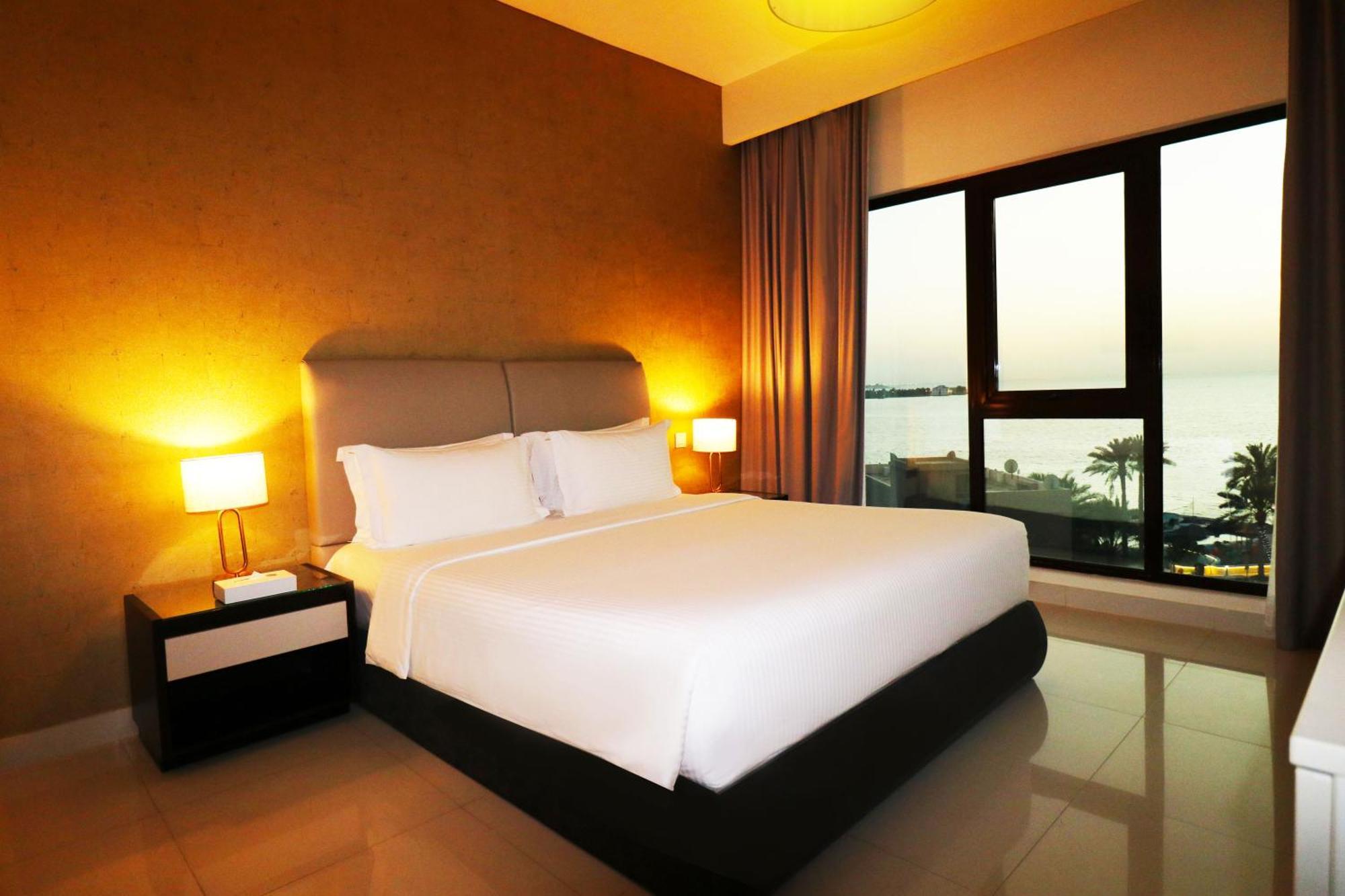 라구나 비치 럭셔리 리조트 앤 스파 마나마 외부 사진 The photo shows a modern hotel bedroom with a large, comfortable bed dressed in white linens. The room features warm lighting, including bedside lamps, creating a cozy atmosphere. There are large windows with a view of the outdoors, possibly overlook